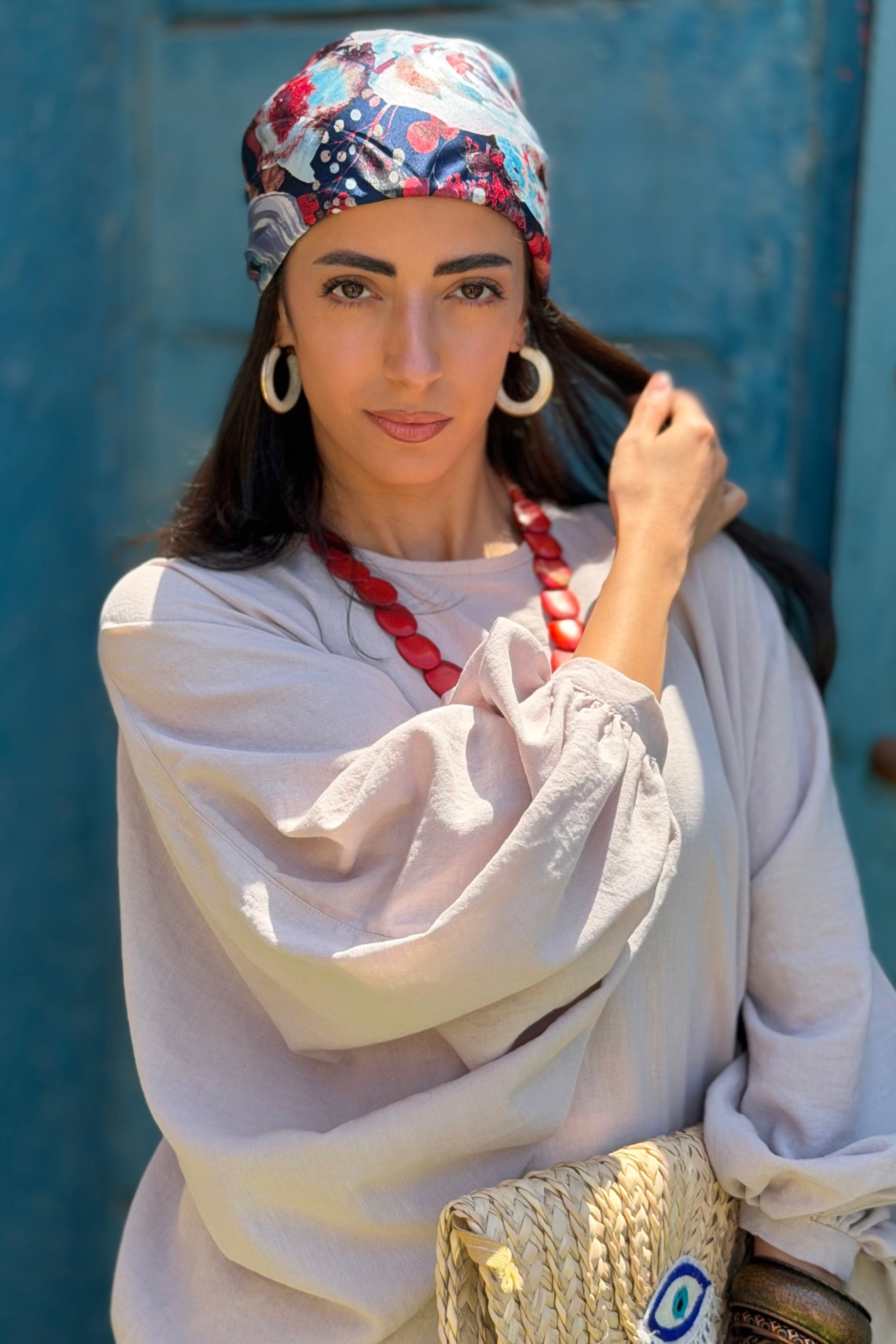 A woman wearing a light beige linen tunic, a colorful patterned headscarf, and a red beaded necklace, holding a woven straw clutch bag with an evil eye design, set against a rustic blue wooden door.